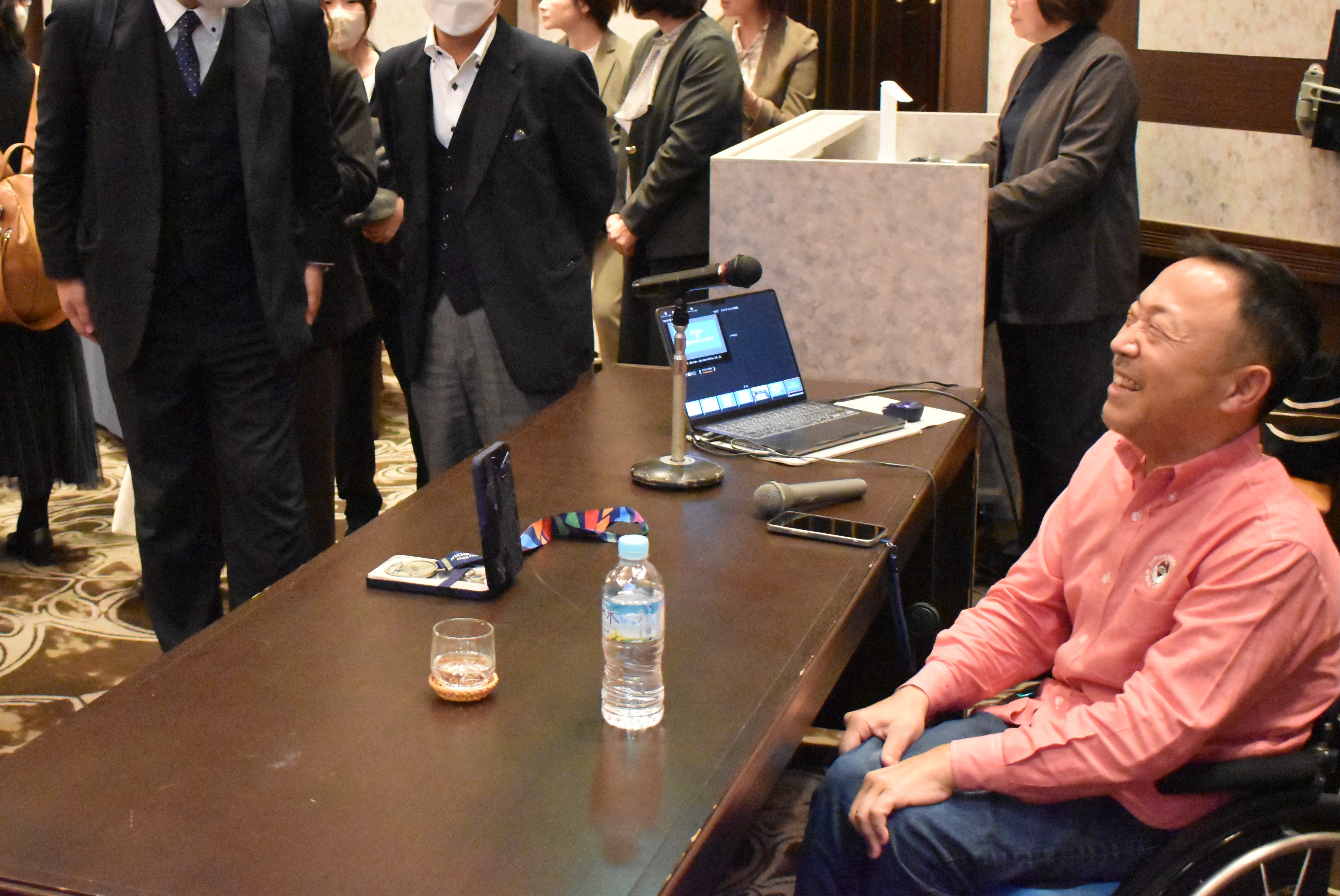 写真：講演会場の卓上にノートPCとメダルが並び、参加者が周囲で立ち話をしている様子