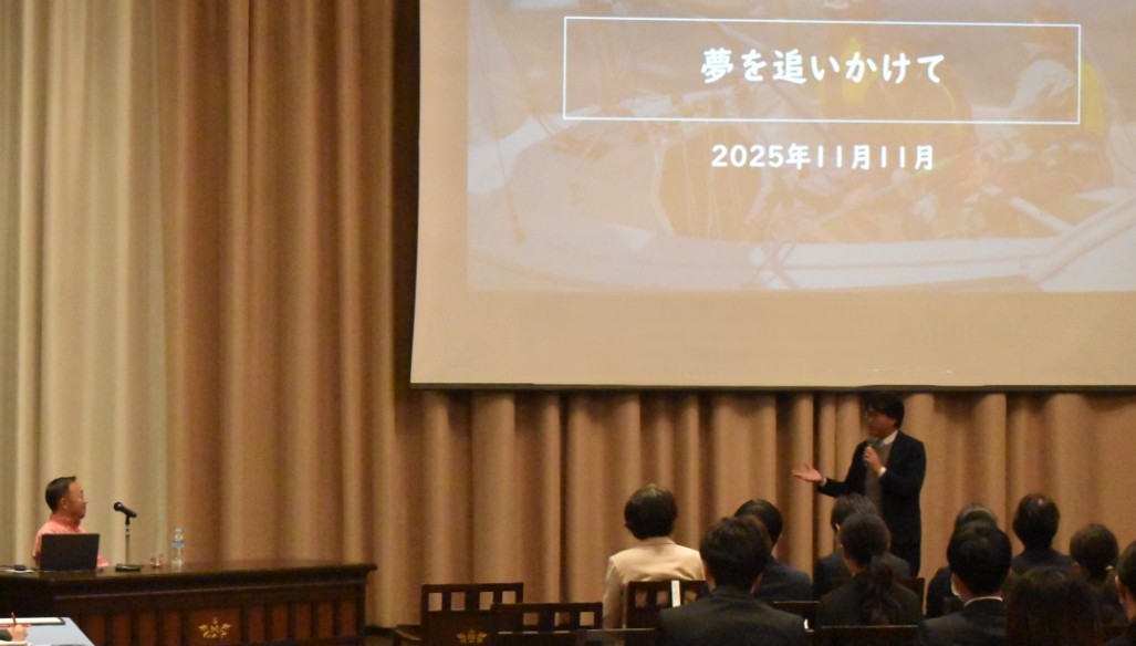 写真：講演会場で登壇者がマイクを手に聴衆へ向けて話しており、前方スクリーンには「夢を追いかけて2025年11月11日」のタイトルが表示されている。